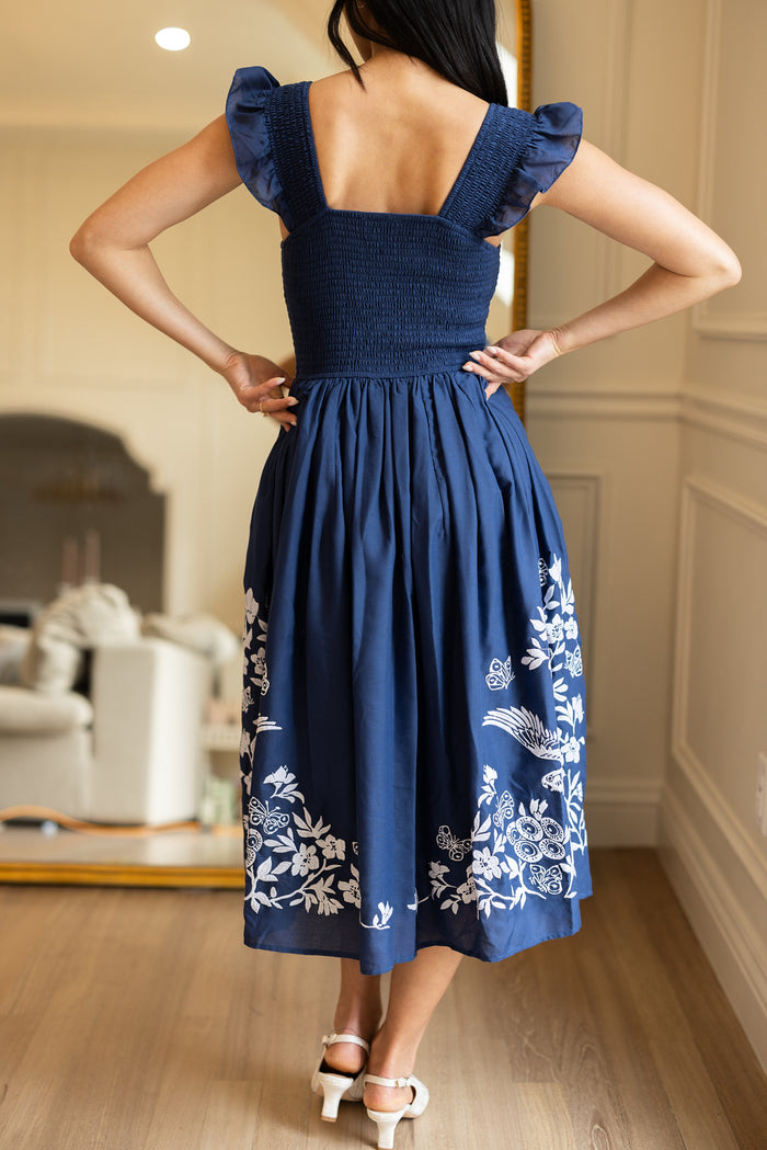 A woman stands indoors with her back to the camera, wearing the Ivy City Co Finch Dress in Blue—featuring a smocked bodice, ruffled sleeves, and white embroidery near the hem—paired with white heeled sandals.