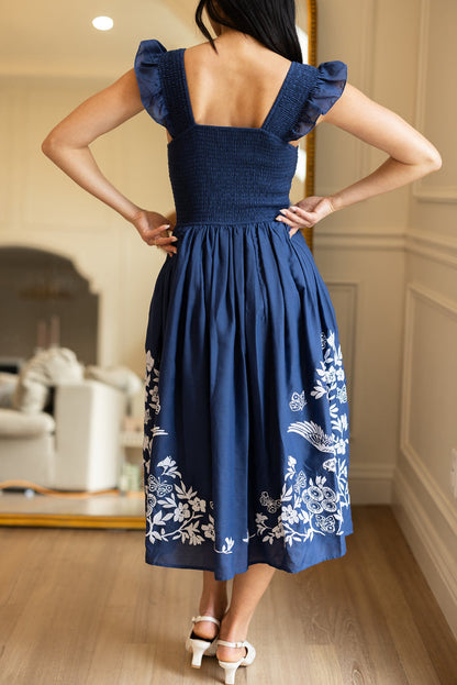 A woman stands indoors with her back to the camera, wearing the Ivy City Co Finch Dress in Blue—featuring a smocked bodice, ruffled sleeves, and white embroidery near the hem—paired with white heeled sandals.
