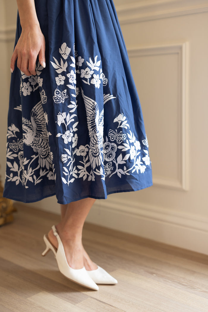 A person stands indoors on a wooden floor, showing only their lower half and white heeled shoes while wearing the Ivy City Co Finch Dress in Blue.