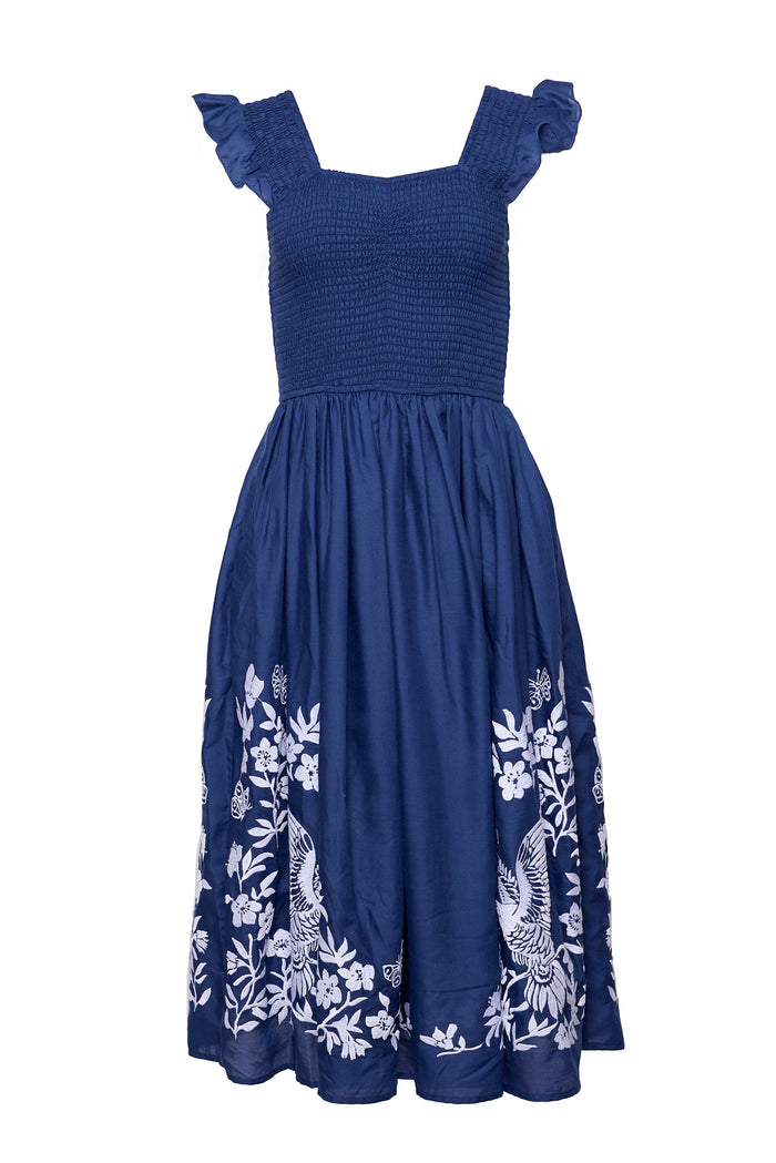 The Finch Dress in Blue by Ivy City Co is a sleeveless cotton dress with ruffled straps, a smocked bodice, and a flared skirt adorned with white floral and bird embroidery along the hem.
