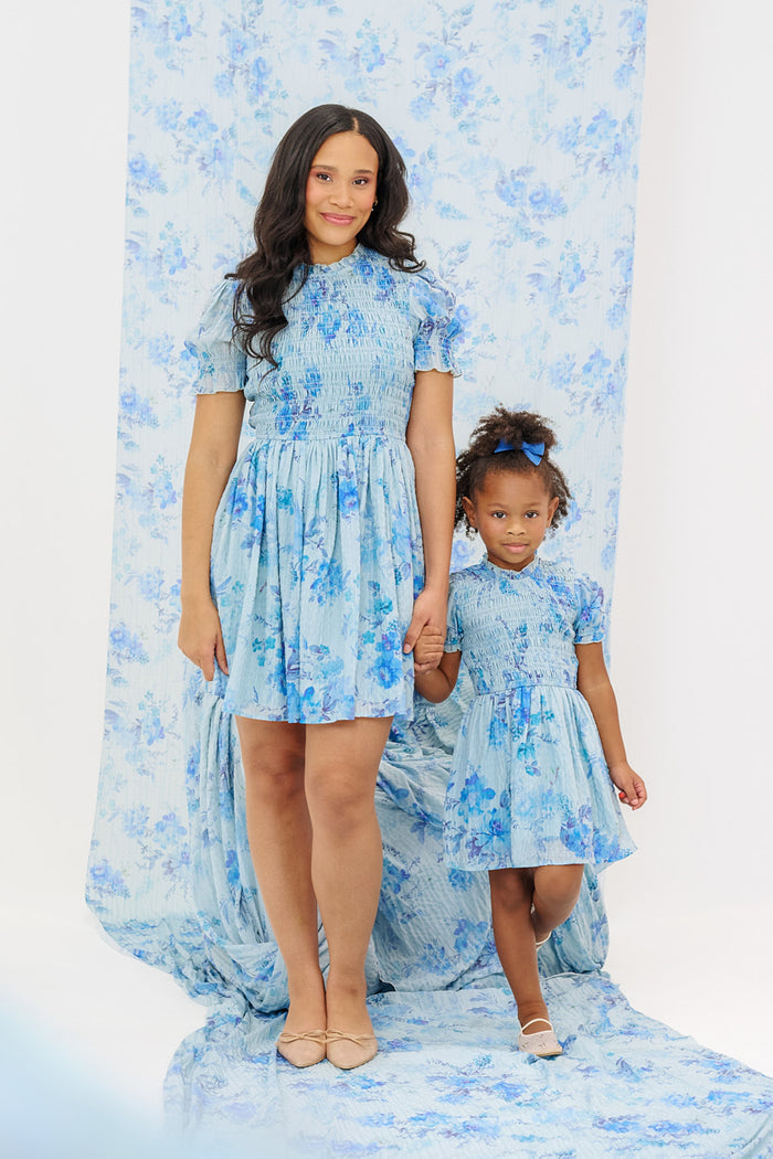 A woman and a young girl hold hands in matching Ivy City Co Flynn Dress in Aquarelle Fleur, each featuring smocked bodices. The blue floral background complements their coordinated look as they smile at the camera.