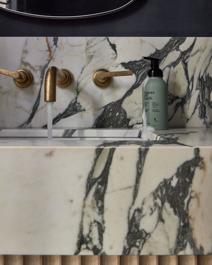 Guests on Earth Foaming Hand Soap on marble sink ledge beside brass tap with running water and a drip of foam
