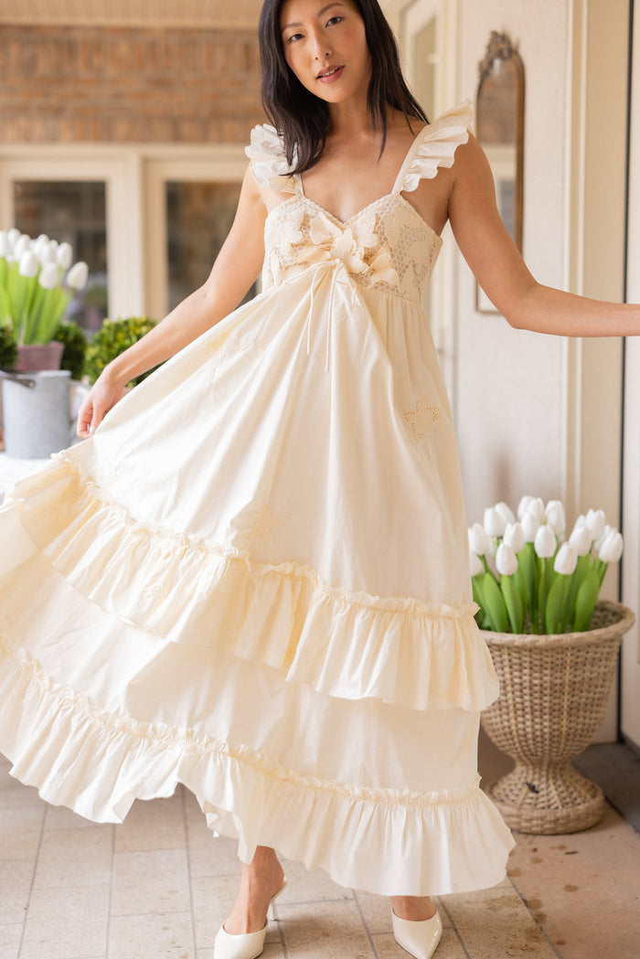 A woman poses indoors in the Ivy City Co Gaia Butterfly Dress in Ivory, a ruffled, sleeveless maternity-friendly spring dress with lace details. White tulips in baskets and pots decorate the background as she stands on a tiled floor.