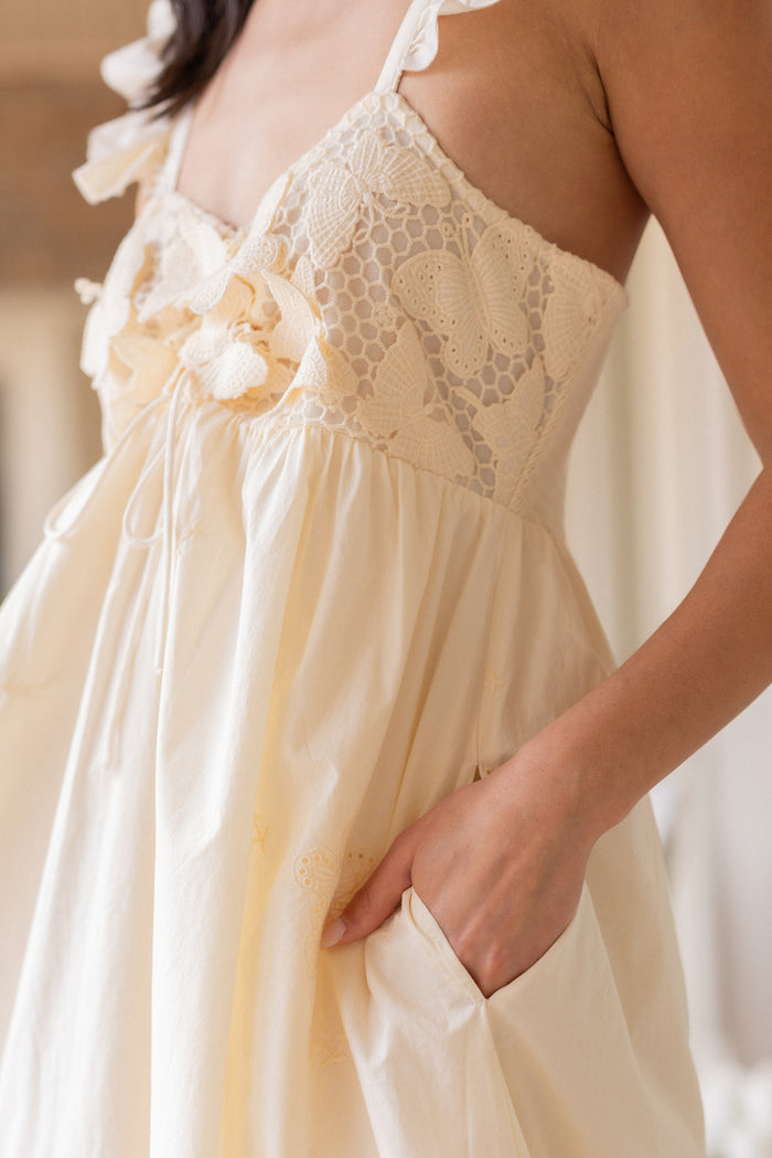 A woman wears the Gaia Butterfly Dress in Ivory by Ivy City Co—an airy, cream-colored maxi with lace, butterfly embroidery, ruffled straps, and a maternity-friendly fit. One hand rests in her pocket as the light fabric flows gracefully.