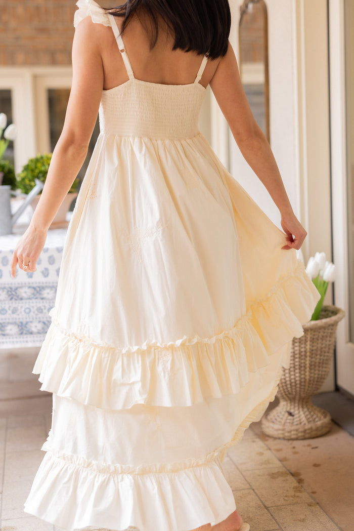 A woman with dark hair wears the Ivy City Co Gaia Butterfly Dress in Ivory, holding up one side of the sleeveless, ruffled maxi skirt. She stands indoors near a table with a blue patterned cloth and white tulips.