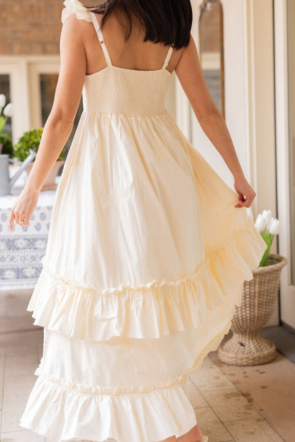 A woman with dark hair wears the Ivy City Co Gaia Butterfly Dress in Ivory, holding up one side of the sleeveless, ruffled maxi skirt. She stands indoors near a table with a blue patterned cloth and white tulips.