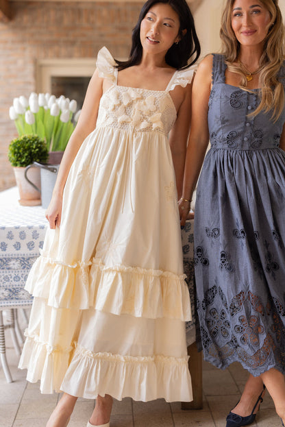 Two women stand indoors by a table with a white floral cloth, white tulips, and a potted shrub. One wears Ivy City Co’s Gaia Butterfly Dress in Ivory—an embroidered, ruffled, maternity-friendly spring style. The other wears a blue dress.