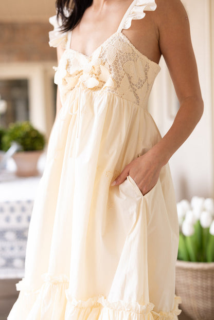 A woman stands indoors wearing the Ivy City Co Gaia Butterfly Dress in Ivory, featuring lace trim, ruffled straps, and a bow detail. With a hand in her pocket and white tulips behind her, she embodies chic style in this maternity-friendly spring look.
