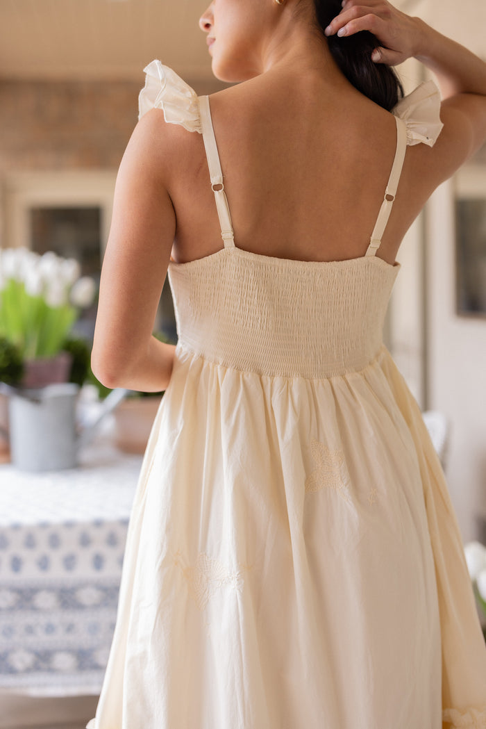 A woman with dark hair, seen from behind, wears the Ivy City Co Gaia Butterfly Dress in Ivory. She touches her hair while a table with a floral tablecloth and a pot of white flowers appears in the background.