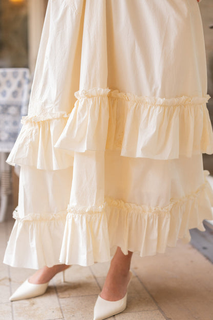 A close-up of the Gaia Butterfly Dress in Ivory by Ivy City Co features tiered, ruffled cream fabric, paired with pointed white heels. The softly blurred background highlights light tile flooring and a glimpse of furniture.