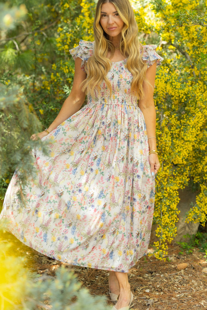 A woman with long blonde hair stands outdoors among green trees and yellow flowers, smiling softly at the camera in the Ivy City Co Hattie Dress in Cordelia Floral, holding out the A-line midi skirt.