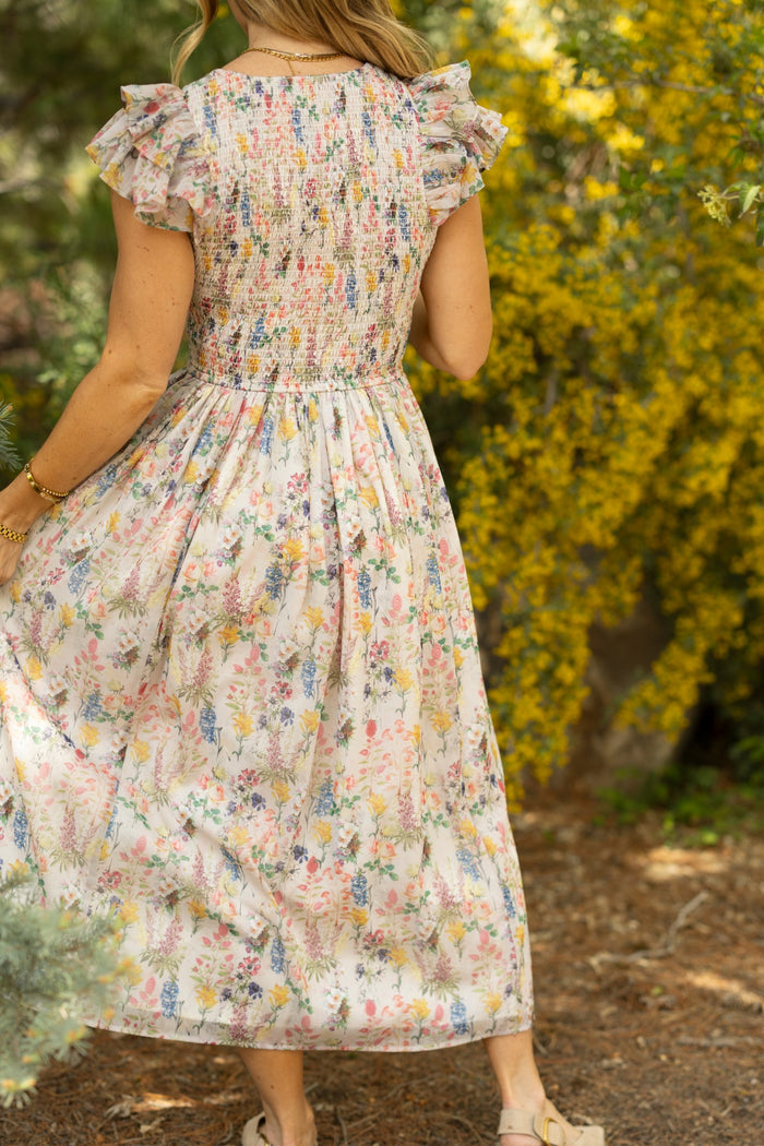 A woman in the Ivy City Co Hattie Dress in Cordelia Floral, an A-line midi with ruffled sleeves, stands outdoors among green foliage and yellow flowers, sunlight highlighting this maternity-friendly look.