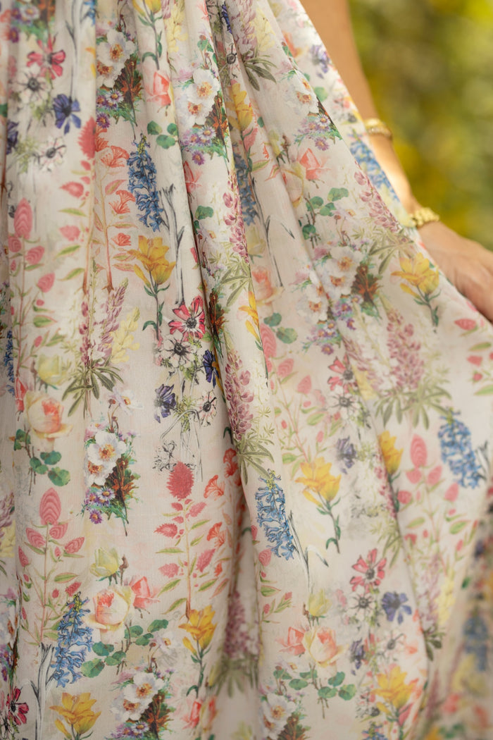 Close-up of Ivy City Co's Hattie Dress in Cordelia Floral—a light-colored, maternity-friendly A-line midi with a vibrant pink, yellow, blue, and green floral print. Gold-braceleted hand gently holds the fabric; background softly blurred.
