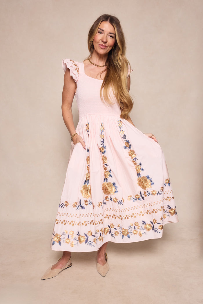 A woman models the Ivy City Co Hattie Dress in Embroidered Spring Floral, featuring ruffled cap sleeves and a maternity-friendly fit, styled with beige flats against a neutral background.