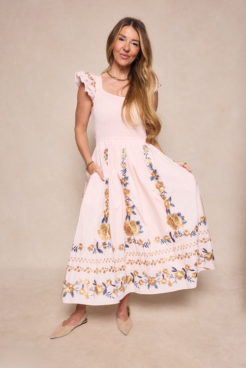 A woman models the Ivy City Co Hattie Dress in Embroidered Spring Floral, featuring ruffled cap sleeves and a maternity-friendly fit, styled with beige flats against a neutral background.