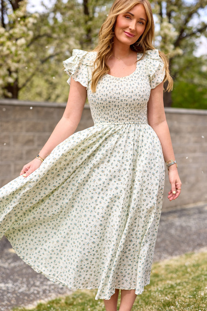 A woman stands on grass, holding the skirt of her Ivy City Co Hattie Dress in Madeline Blue—a white, floral-patterned cotton midi dress with ruffled short sleeves. Trees and a stone wall are behind her on a sunny day.