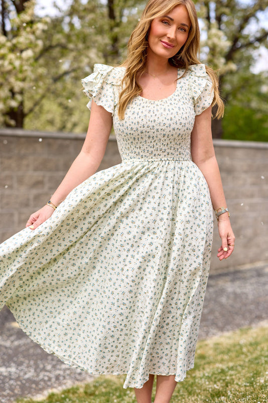 A woman stands on grass, holding the skirt of her Ivy City Co Hattie Dress in Madeline Blue—a white, floral-patterned cotton midi dress with ruffled short sleeves. Trees and a stone wall are behind her on a sunny day.