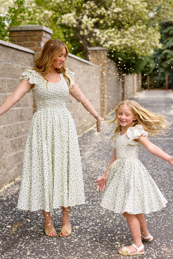 A woman and a young girl smile and play amid falling flower petals, both wearing matching Ivy City Co Hattie Dresses in Madeline Blue on a tree-lined path.