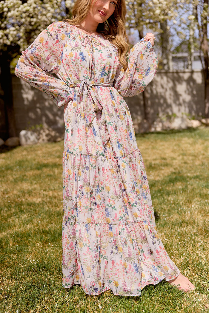 A woman stands barefoot on grass in the Ivy City Co Imogen Dress in Cordelia Floral—a flowy chiffon maxi with billowy sleeves and tied waist—surrounded by blooming trees for an effortlessly elegant, maternity-friendly look.