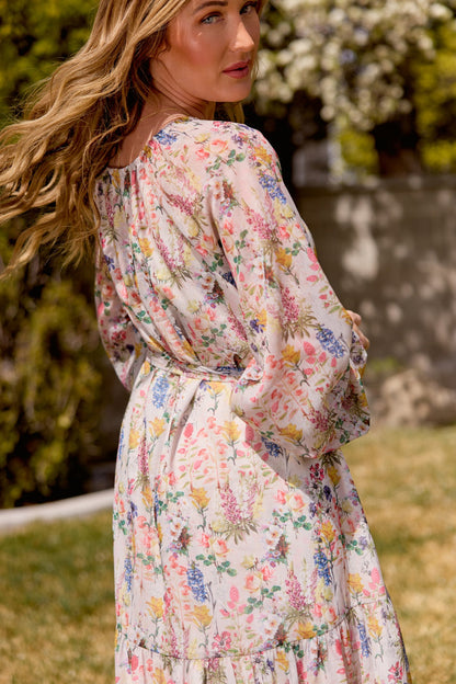 A woman with wavy blonde hair stands outdoors in the Imogen Dress in Cordelia Floral by Ivy City Co, a flowy chiffon maxi in soft pastels. She looks over her shoulder amid blurred greenery and white flowers. This dress is maternity friendly.