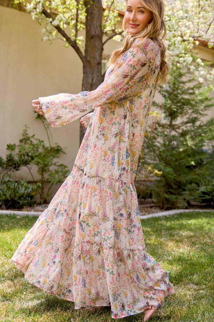 A woman in the Imogen Dress in Cordelia Floral by Ivy City Co, a flowy chiffon maternity-friendly maxi, stands barefoot on grass, smiling and turning sideways in a sunlit garden with trees and shrubs behind her.