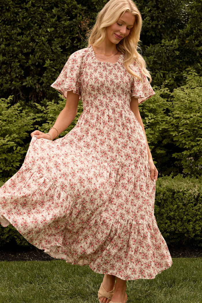 A woman stands on grass in the Ivy City Co Jovie Dress in Ivory Heritage Rose, a maternity and nursing-friendly dress with pink floral patterns. She smiles, holding out one side of her flowing dress amid lush green bushes.