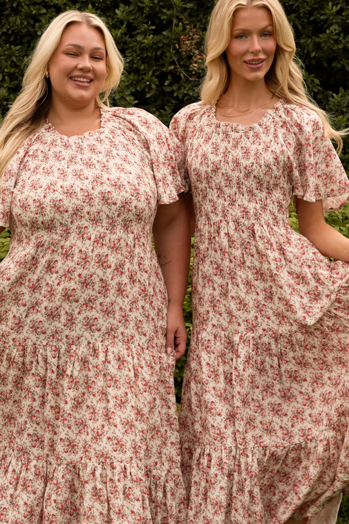 Two women smile outdoors in matching Ivy City Co Jovie Dresses in Ivory Heritage Rose—long, short-sleeved, floral, and maternity- and nursing-friendly—standing together before lush green shrubbery.