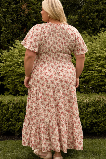 A person with blonde hair stands outdoors with their back to the camera, surrounded by greenery, wearing the maternity and nursing-friendly Jovie Dress in Ivory Heritage Rose by Ivy City Co.