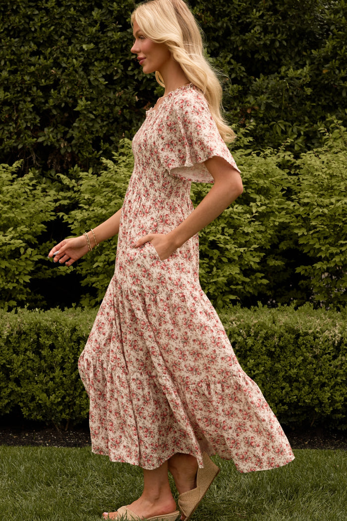 A woman with long blonde hair walks on grass, wearing the Ivy City Co Jovie Dress in Ivory Heritage Rose—maternity and nursing friendly—featuring pink and red florals, paired with beige wedge sandals. Lush green bushes are in the background.