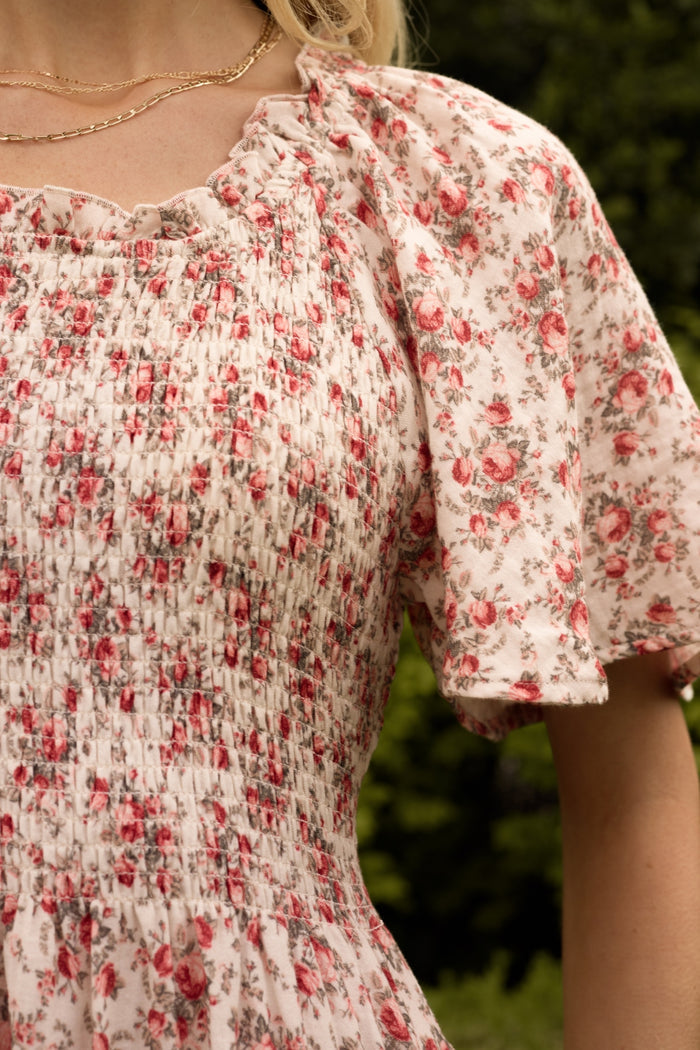 A close-up of a person wearing the Ivy City Co Jovie Dress in Ivory Heritage Rose, which features a smocked bodice and short puff sleeves. Maternity and nursing friendly, it’s ideal for matching mom and me moments with blurred greenery behind.
