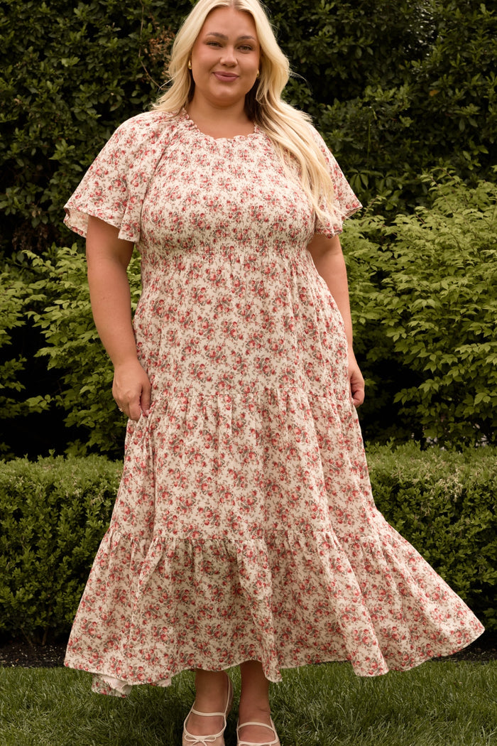 A woman with long blonde hair stands outdoors by green bushes, smiling slightly, wearing the Ivy City Co Jovie Dress in Ivory Heritage Rose—designed to be both maternity and nursing friendly.