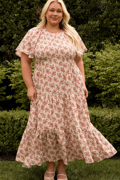 A woman with long blonde hair stands outdoors by green bushes, smiling slightly, wearing the Ivy City Co Jovie Dress in Ivory Heritage Rose—designed to be both maternity and nursing friendly.