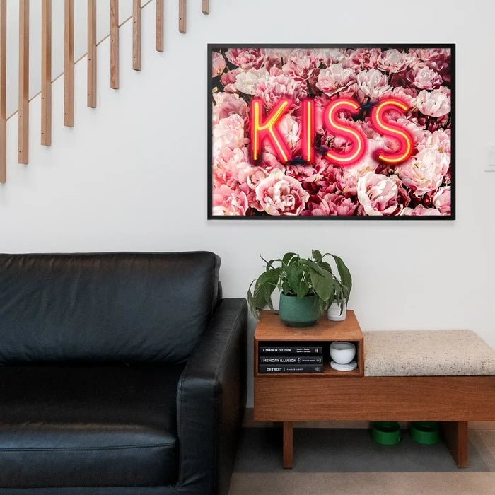 Framed wall art featuring a glowing neon KISS sign layered over a vibrant background of pink roses in a modern living room.