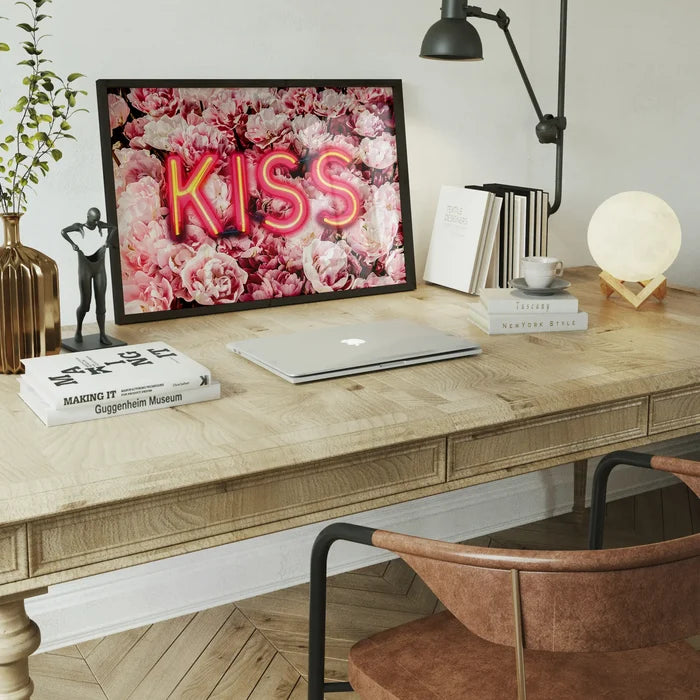 Framed print featuring a bright neon KISS sign glowing over a vibrant background of pink roses on a wooden desk.