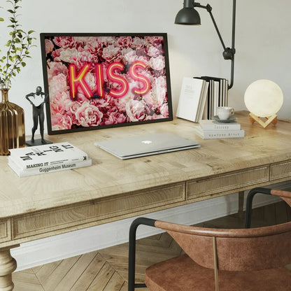 Framed print featuring a bright neon KISS sign glowing over a vibrant background of pink roses on a wooden desk.