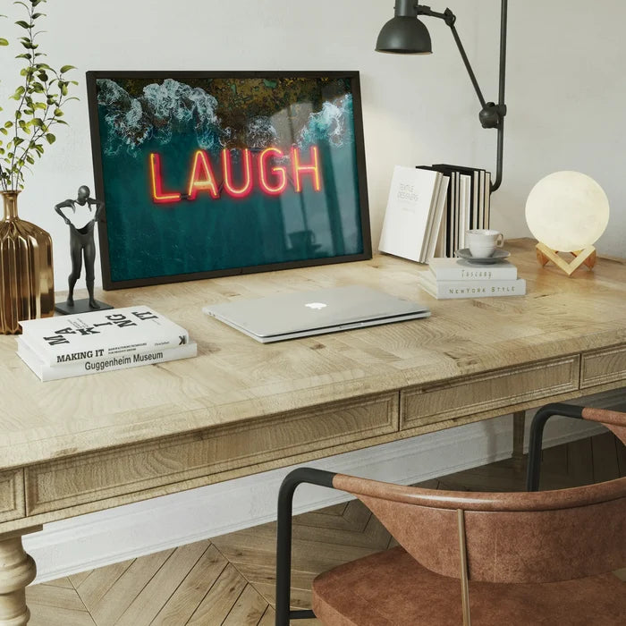 Glossy framed print featuring a bright red LAUGH neon sign overlaying a vibrant ocean wave scene on a wooden desk.
