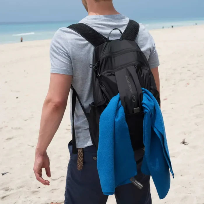 Person on a beach wearing a Lifeventure Packable Backpack 25 Litres in black with a blue towel fed through it