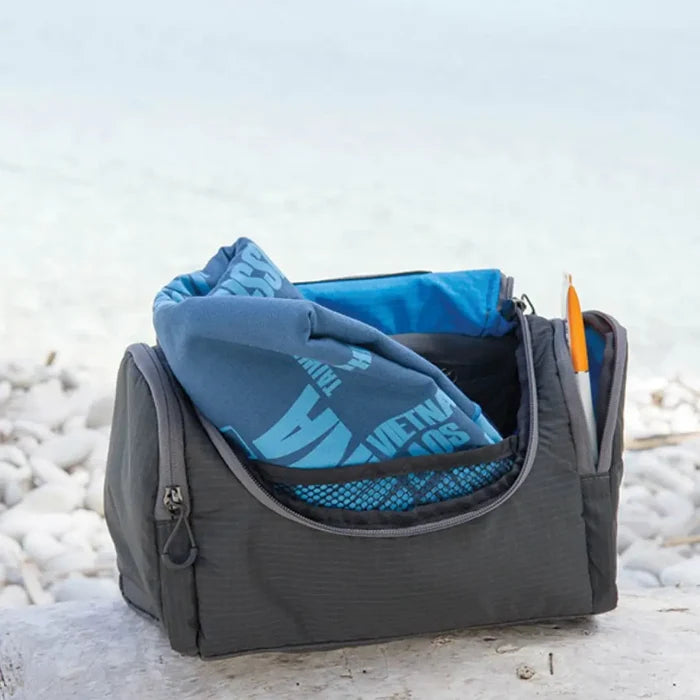 Lifeventure Travel Wash Holdall Grey shown outdoors on a beach with a travel towel protruding from the main compartment