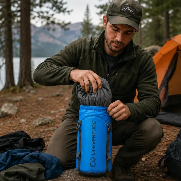 Man stuffing a jacket into a Lifeventure Ultralight Compression Sack 10 Litres in blue whilst camping in the woods by a lake