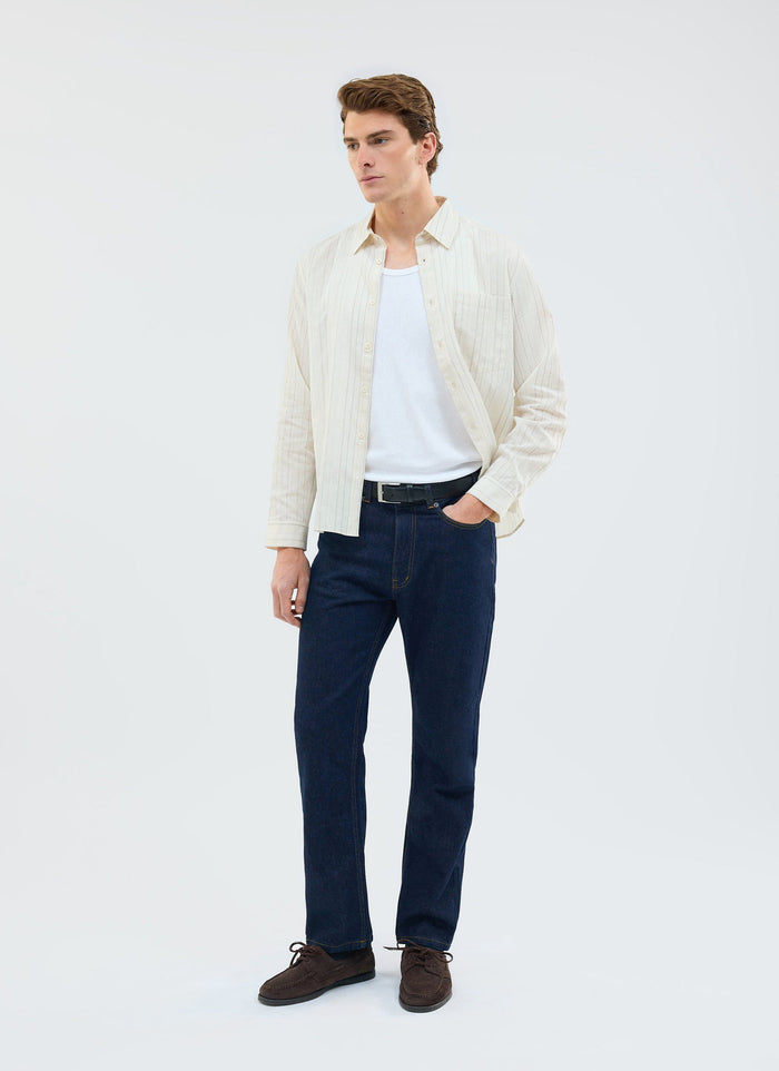 A model wears the ecru Lorenzo Pearce Shirt by Percival, styled open over a white vest and dark blue jeans.