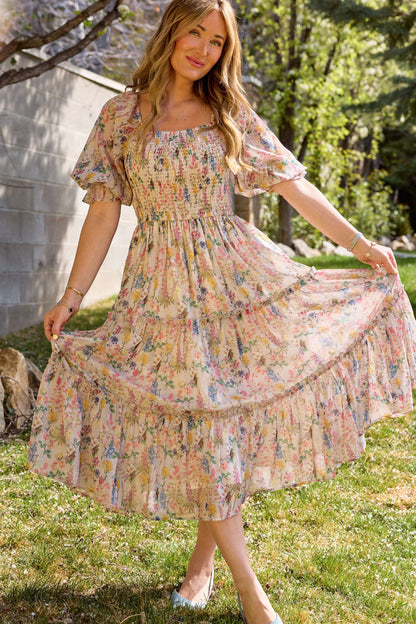 A woman stands on grass, holding out her Ivy City Co Madeline Dress in Cordelia Floral with ruffled sleeves and hem. Sunlight filters through the trees, highlighting her A-line midi silhouette against a stone wall backdrop.