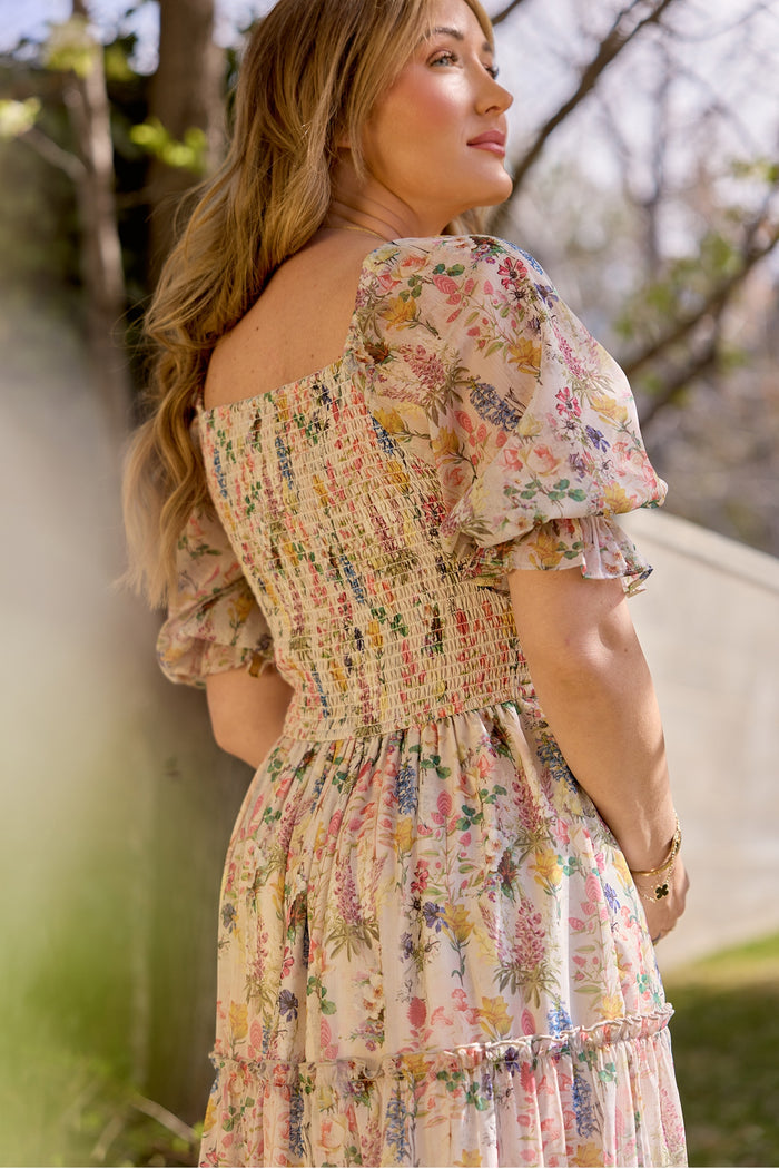 A woman stands outdoors in Ivy City Co’s Madeline Dress in Cordelia Floral, featuring puffed short sleeves and a smocked bodice. Sunlight highlights her long, wavy hair as she looks to the side, surrounded by greenery and trees.