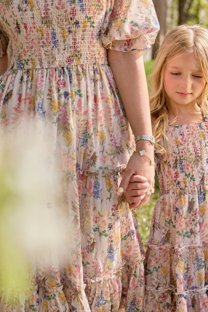 A woman and young girl hold hands outdoors, both wearing matching Ivy City Co Madeline Dress in Cordelia Floral. Their partially visible faces draw attention to their clasped hands and the elegant floral dress details.