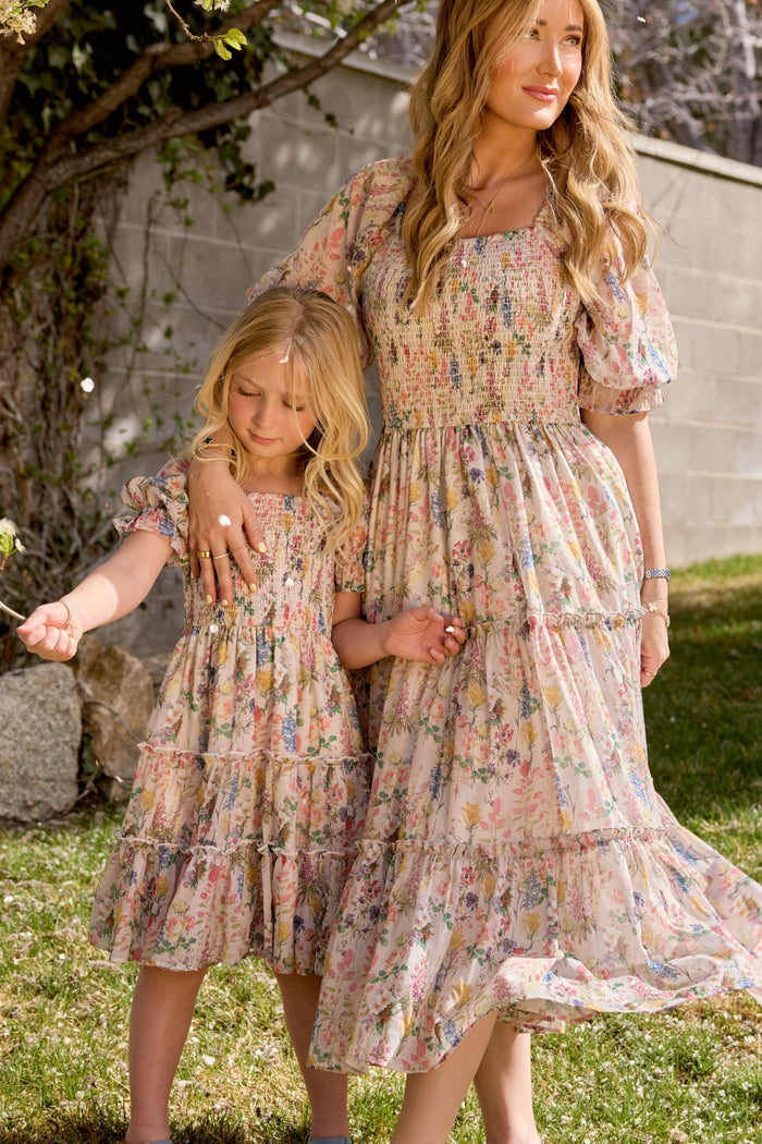 A woman and a young girl stand on grass, holding hands in matching Ivy City Co Madeline Dresses in Cordelia Floral. Their light A-line midi dresses with puff sleeves and smocked bodices glow as sunlight highlights their blond hair.