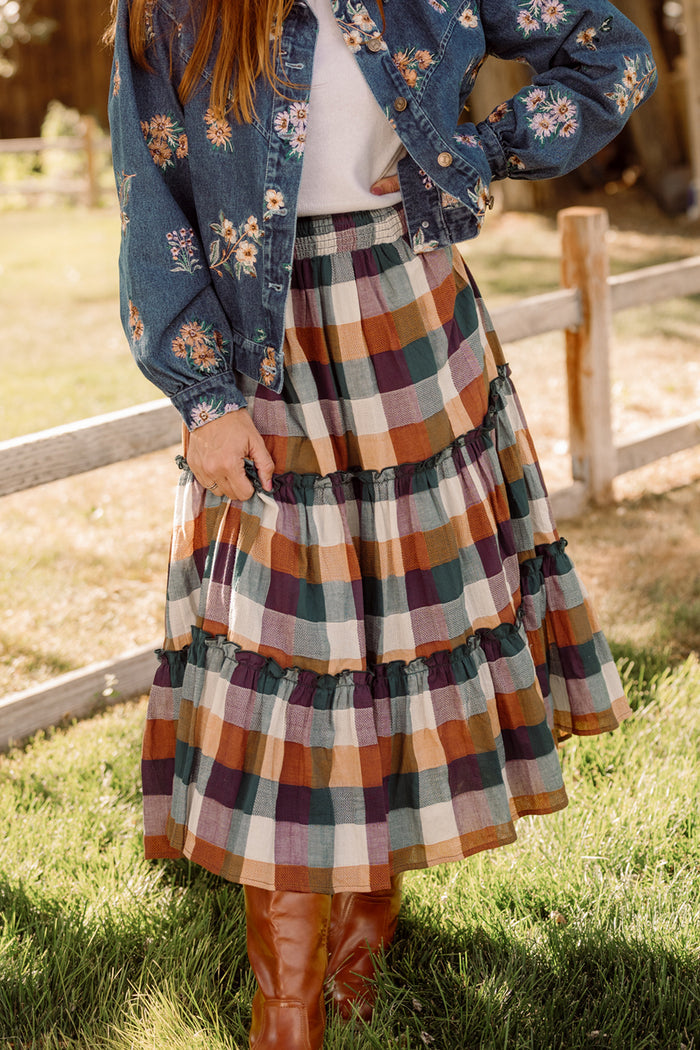 A person wearing a floral denim jacket, a white top, and the Ivy City Co Madeline Skirt in Maize Gingham—a tiered plaid cotton-blend skirt featuring autumn colors and convenient pockets—stands on green grass near a wooden fence, completing the look with brown knee-high boots.