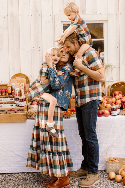A family of four poses in front of a white wall and farm stand with apples. The mother and two children wear matching plaid outfits, with the mother’s look completed by the stylish Madeline Skirt in Maize Gingham from Ivy City Co. The father wears a plaid shirt and jeans. The family hugs and laughs together.