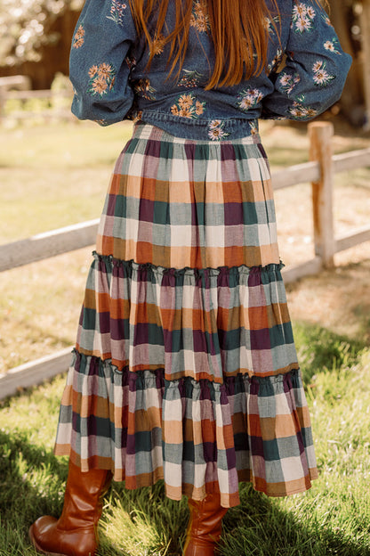A woman with long hair stands on grass near a wooden fence, wearing a floral denim shirt, tall brown boots, and the Madeline Skirt in Maize Gingham by Ivy City Co. This cotton-blend skirt features pockets, combining comfort and practicality with its vibrant, playful design.
