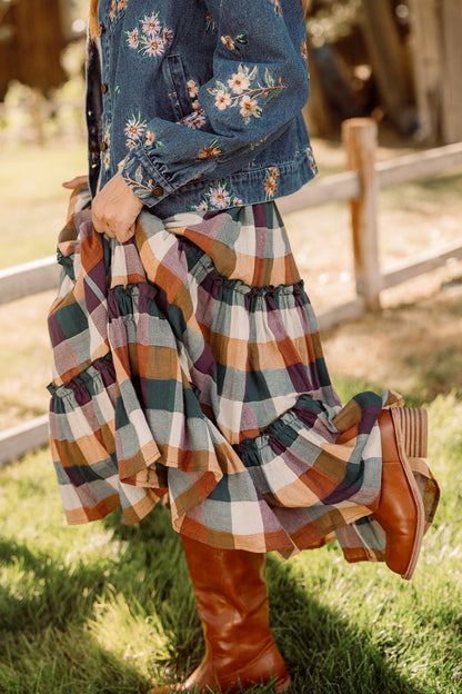 A person wearing a floral denim jacket, the Ivy City Co Madeline Skirt in Maize Gingham with pockets, and brown heeled boots stands on grass near a wooden fence, holding the skirt up slightly.