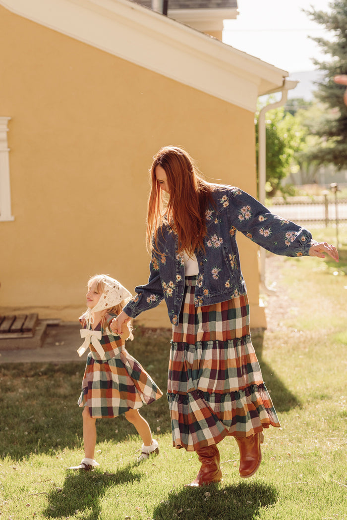 A woman and a young girl, both in plaid dresses, walk hand in hand across the grass before a yellow house. The woman is wearing boots and a floral jacket paired with the Ivy City Co Madeline Skirt in Maize Gingham, while the young girl wears a bonnet. Sunlight gently illuminates them.