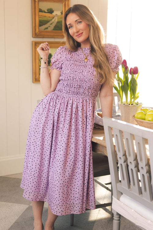 A woman in the Marcell Dress in Purple Polka Dot by Ivy City Co leans against a dining table with pink tulips, lemons, and sunlight streaming through the window in a cozy, decorated room.